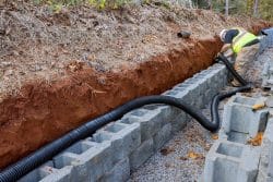A worker in a safety vest installs a black drainage pipe behind a retaining wall made of concrete blocks in a trench, showcasing expert installation of Drainage Systems in Knoxville.