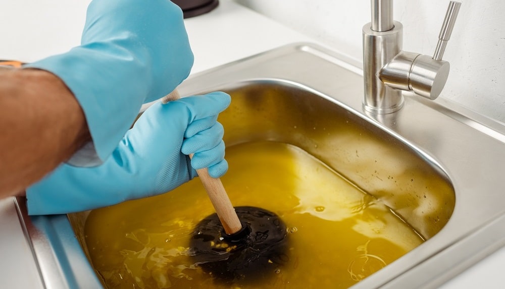 A person wearing blue gloves uses a plunger to unclog kitchen sink drains filled with yellowish water.