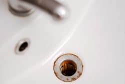 Close-up of a white sink with a metal drain showing visible rust and discoloration around the edge, a common sign that may require unclogging bathroom sink drain solutions.