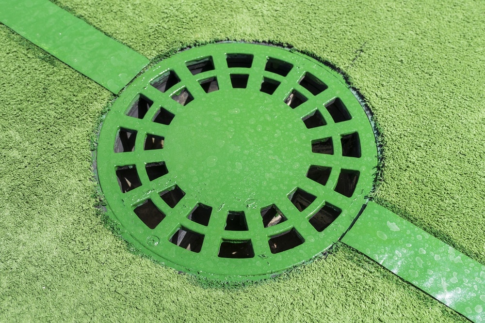 A close-up of a green circular drainage grate set into a green textured surface, intersected by two green strips—an ideal spot for outdoor drain unclogging.
