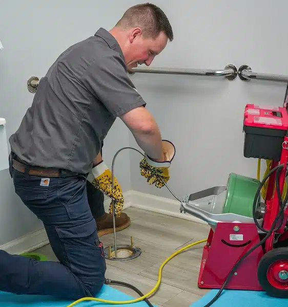 A person kneels on the floor feeding a plumbing snake into a drain, using protective gloves, with equipment nearby.