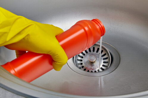 Person's hand in a yellow rubber glove pours pipe cleaner down the drain of a metal kitchen sink. Prevention and cleaning of sewer blockages