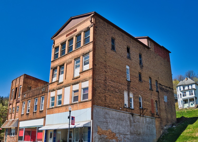 Three-story brick building with boarded-up windows and storefronts at street level, situated on a sloped street under a clear blue sky.
