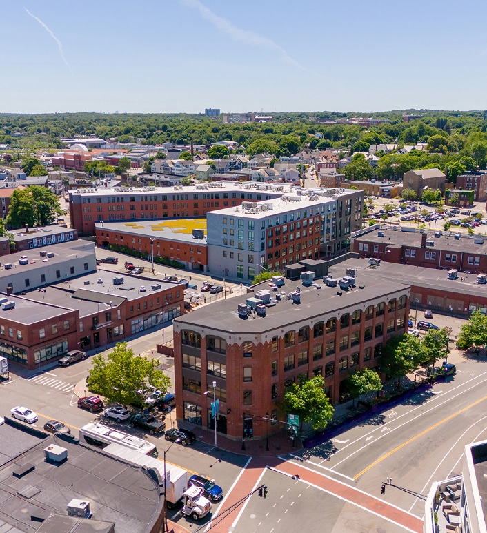 Aerial view of a small city area with brick buildings, streets, and some trees on a sunny day; green landscape visible in the background.