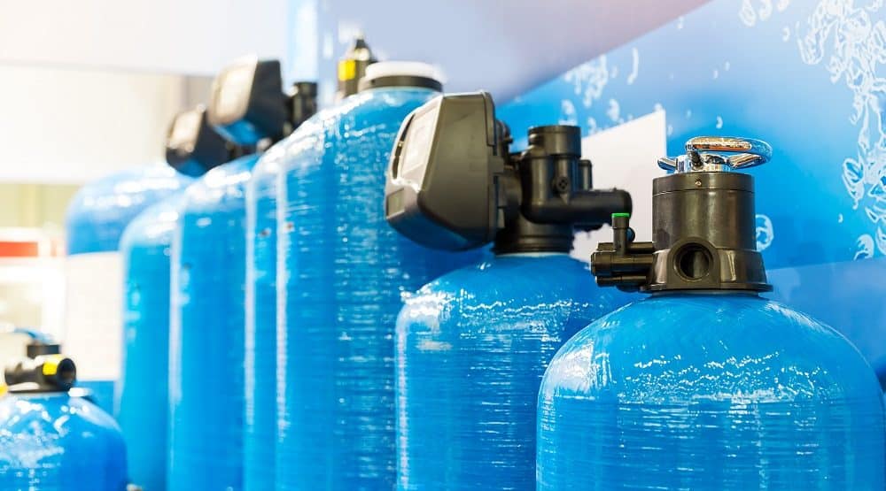 Several large blue industrial water filter tanks with control valves are lined up against a wall with a blue background, showcasing advanced Water Treatment Systems Knoxville trusts for reliable performance.