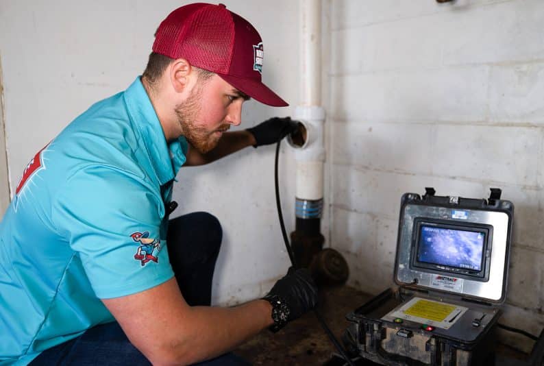 A technician in a blue shirt and red cap inspects a pipe with a camera, viewing the footage on a portable monitor in a utility area.