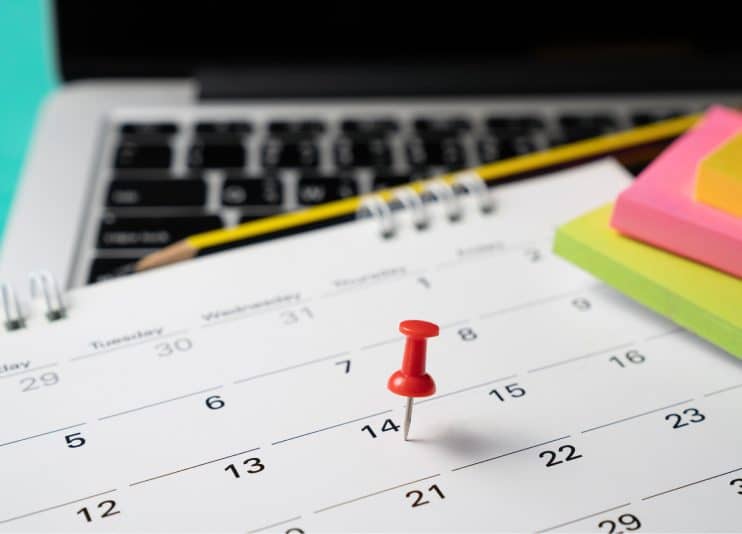 A red pushpin marks a date on a calendar placed on a laptop keyboard, with a pencil and colorful sticky notes nearby.