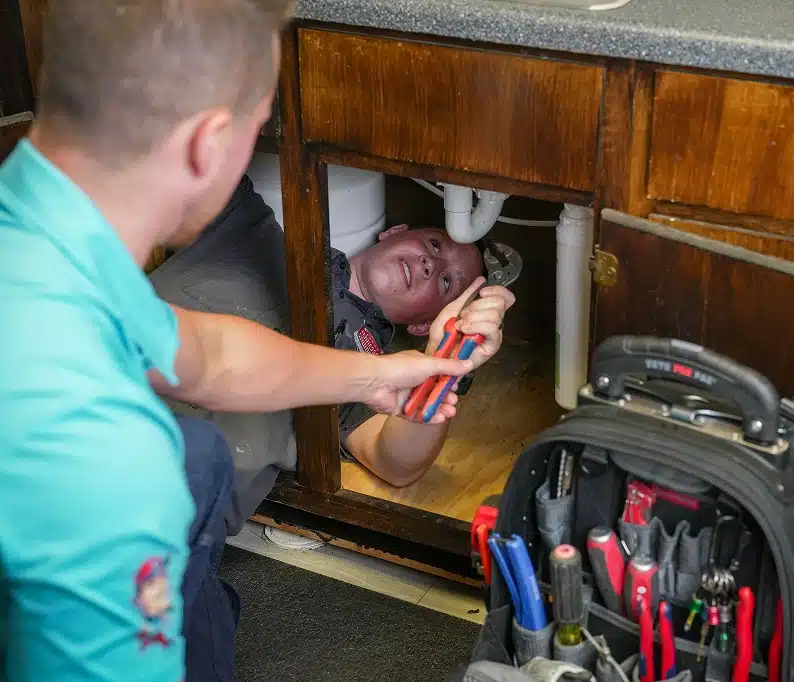 Two plumbers in matching uniforms working under a kitchen sink cabinet, with one lying inside handing tools to the other, alongside a professional tool bag.