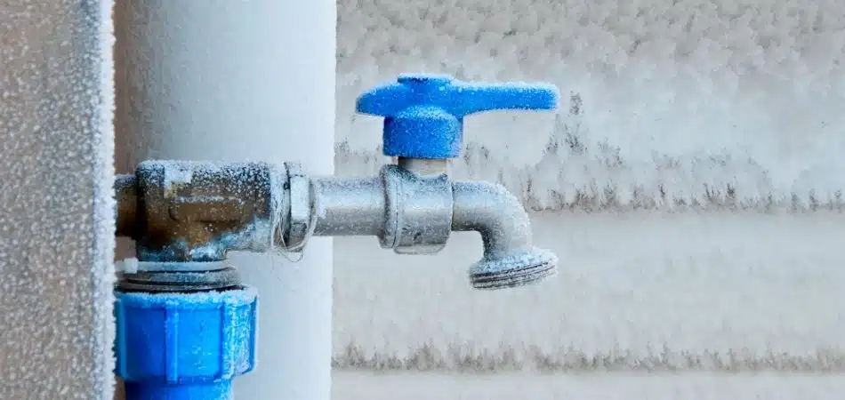 A metal outdoor water faucet with a blue handle is covered in frost and ice, indicating freezing temperatures. Frost is also visible on the surrounding surfaces.