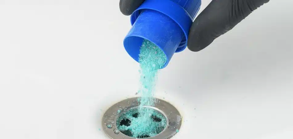 A hand wearing a black glove pours blue drain cleaner powder into a sink drain.