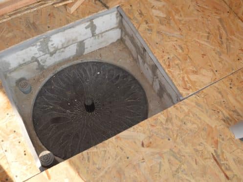 A circular black drain cover, designed to hide sump pump access, is installed in a square cutout within an unfinished oriented strand board (OSB) floor.