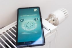 A smartphone displaying a "Water Detected" notification rests on top of a white radiator near a thermostat knob, reflecting 2025 trends in commercial plumbing leak detection technology.