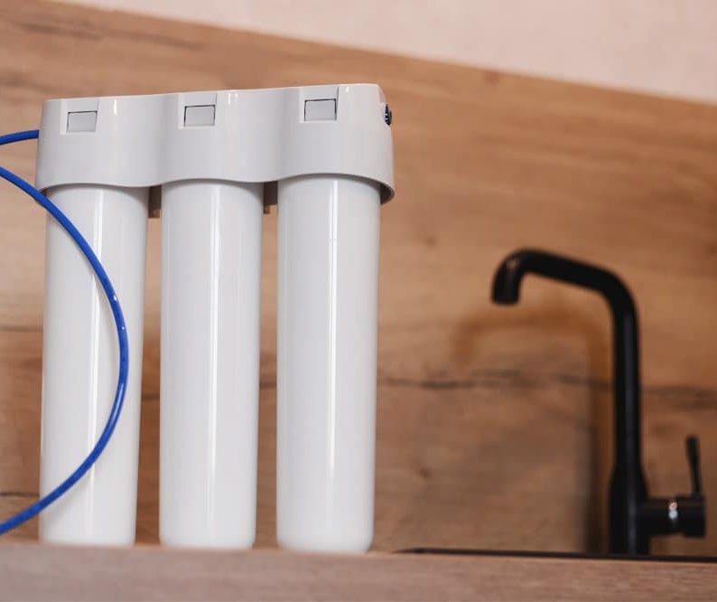 Three-stage water filter system with white cylindrical cartridges and a blue tube, placed on a kitchen countertop near a black faucet—ideal for those seeking reliable Water Treatment Systems in Oak Ridge.