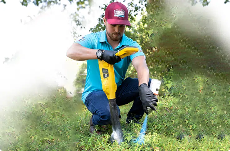 Kelton is wearing gloves and a red cap and uses a handheld yellow underground cable locator while kneeling on grass.