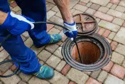 A person wearing gloves and work clothes uses a cable to clean or inspect a drain through an open manhole on a brick-paved surface, demonstrating an effective drain cleaning method.