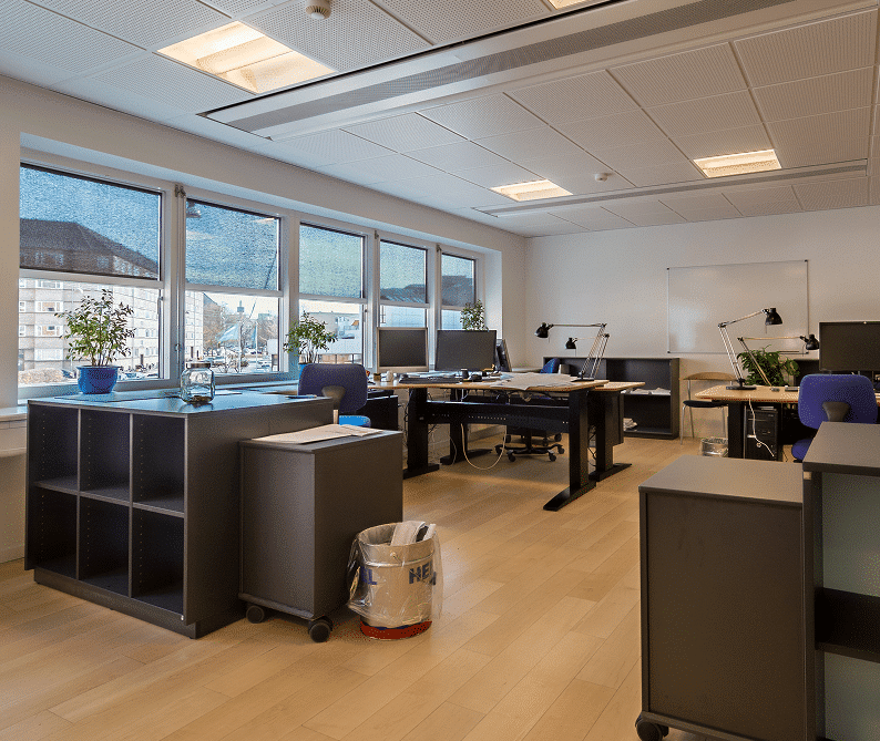 Modern office space with several desks, chairs, computers, small potted plants, large windows, and light wood flooring. The room is well-lit with natural and artificial light.