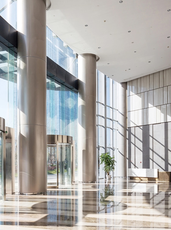 Modern office lobby with tall glass windows, large columns, polished floors, a revolving door, a potted plant, and a white bench. Sunlight casts shadows on the floor and walls.