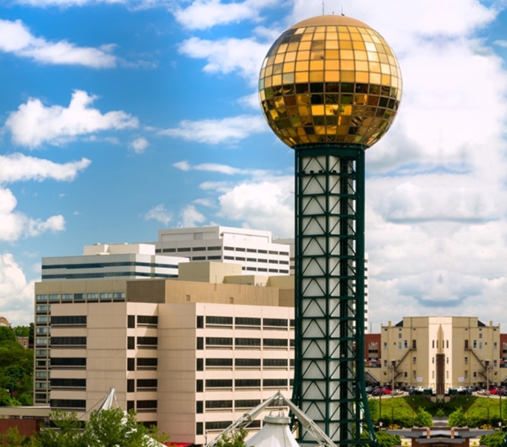 A tall structure with a gold reflective sphere on top stands among office buildings under a partly cloudy sky.