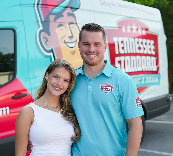 A woman in a white dress and a man in a blue polo shirt stand in front of a van with