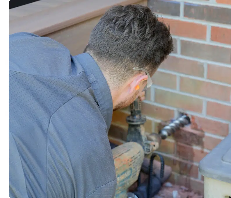 A person wearing safety glasses inspects or repairs an outdoor pipe or valve attached to a brick wall.