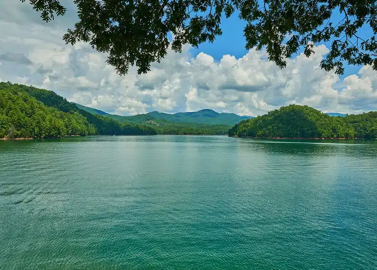 Serene lake with turquoise water, framed by lush green hills under a bright blue sky filled with fluffy clouds in Lenoir City