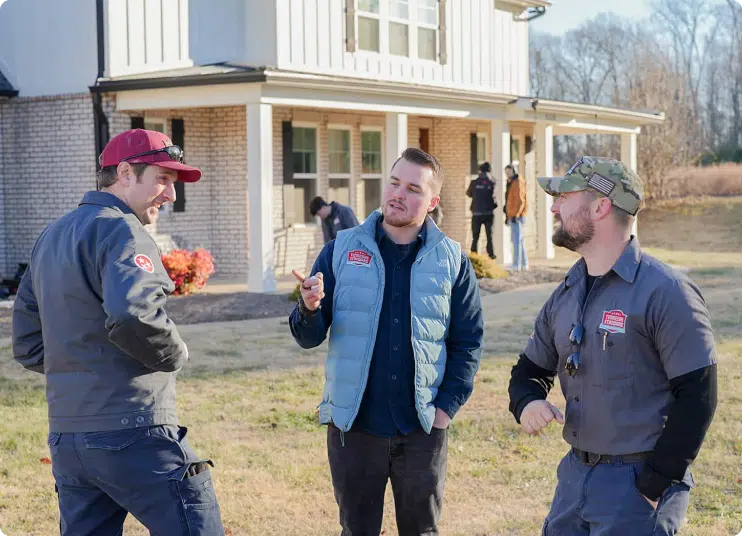 Three men stand outside a house having a conversation, with two wearing work uniforms and one in a light blue vest; others are seen in the background near the house.