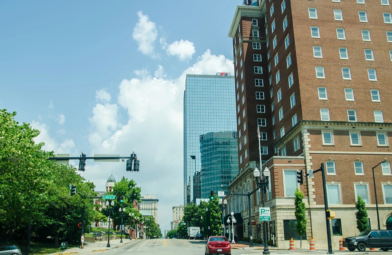 A city street with traffic lights, parked and moving cars, trees, a glass skyscraper, and a brown brick building on a sunny day.