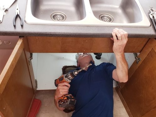 Technician performing kitchen sink drain installation under a double sink using a power drill.