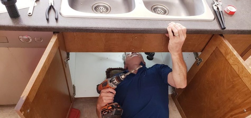 Technician performing kitchen sink drain installation under a double sink using a power drill.