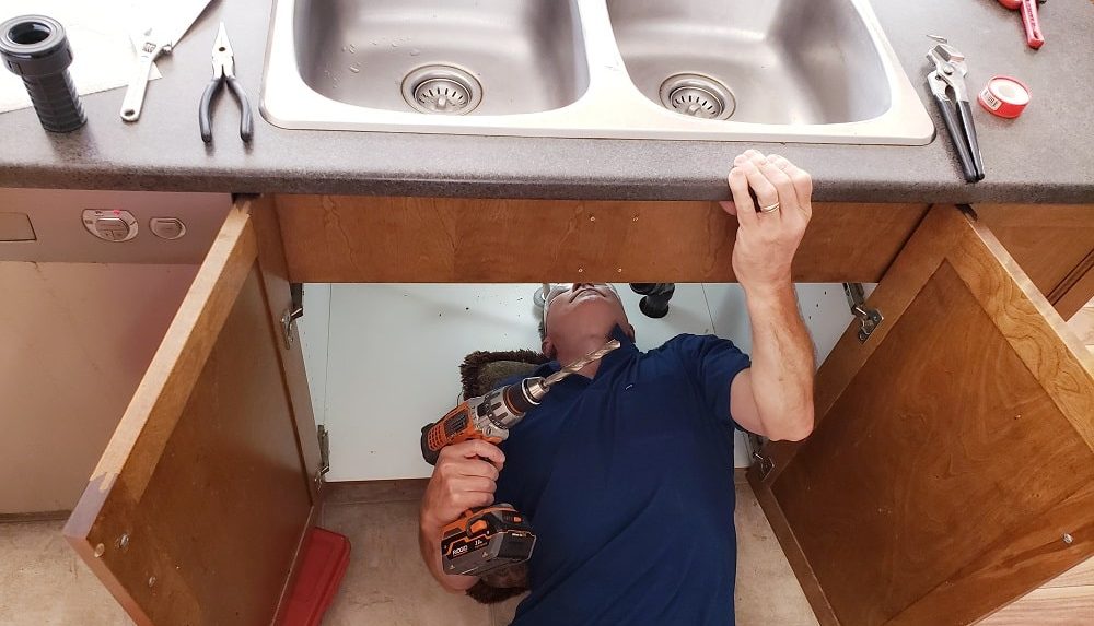 Technician performing kitchen sink drain installation under a double sink using a power drill.