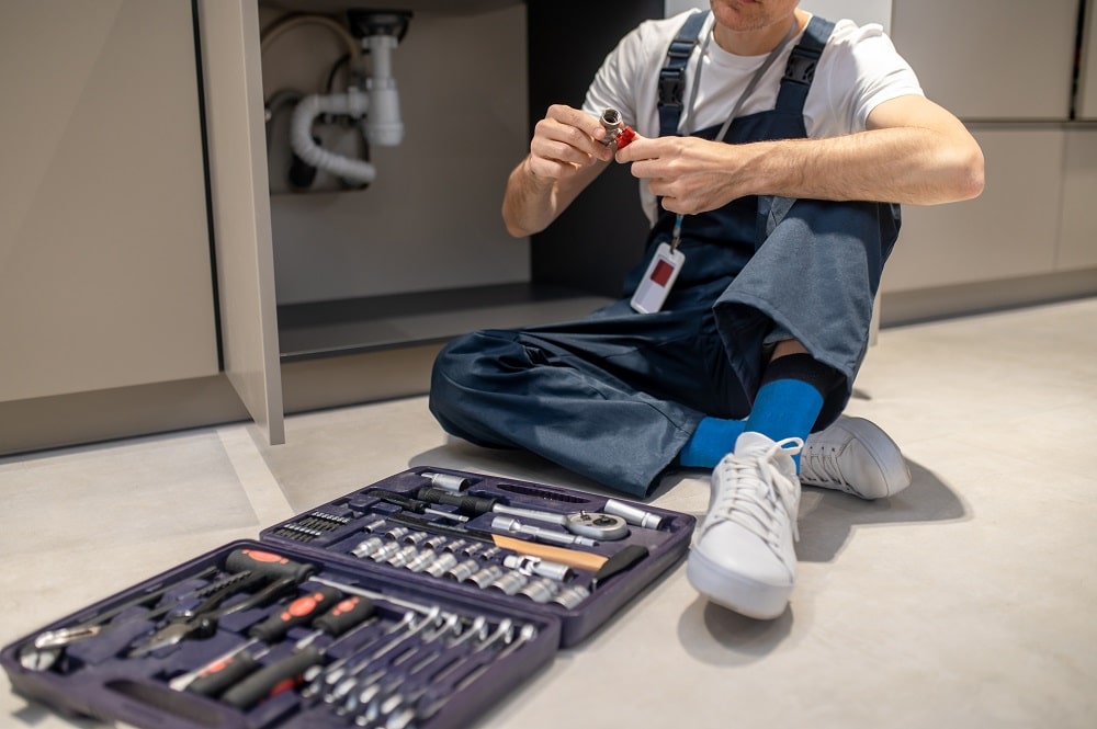 A person in overalls sits on the floor assembling a small plumbing part, with an open toolkit containing various tools nearby—demonstrating practical plumbing maintenance tips in action.