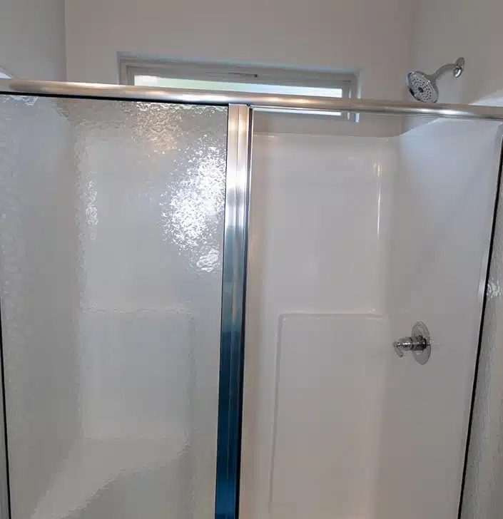 Glass shower with sliding door, chrome frame, and white fiberglass walls; showerhead and control handle visible on the right.