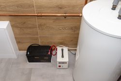 A battery, control unit, and white water heater in Oak Ridge are installed in a room with a tiled floor and wood-patterned wall, featuring visible copper piping.