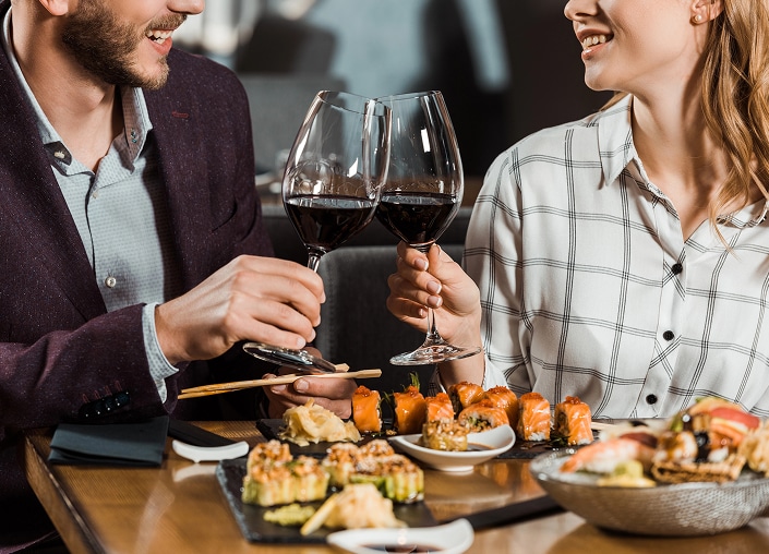 A man and woman clink wine glasses at a restaurant table with assorted sushi and other dishes.