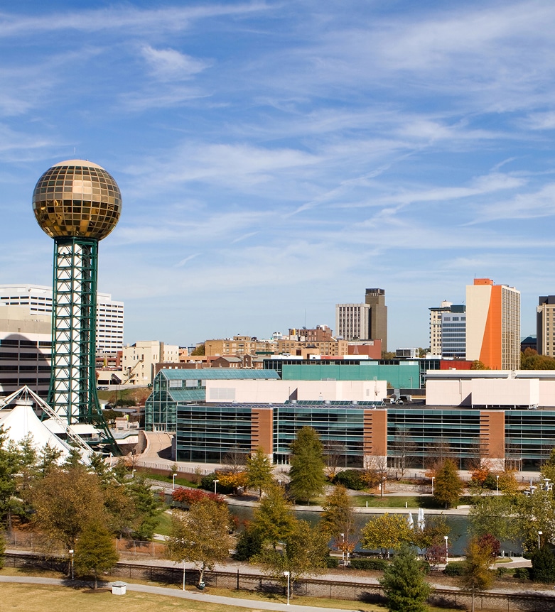 Skyline view of Knoxville, Tennessee, featuring the Sunsphere, modern buildings, and trees under a blue sky.