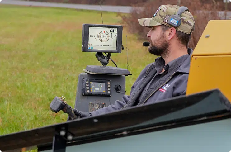 A man in a camo cap and headset operates machinery while monitoring a screen displaying technical data in an outdoor grassy area.