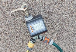 A digital water timer, reflecting 2025 trends in commercial plumbing, is attached to an outdoor faucet with a key in the tap and two hoses connected, all mounted against a speckled wall.