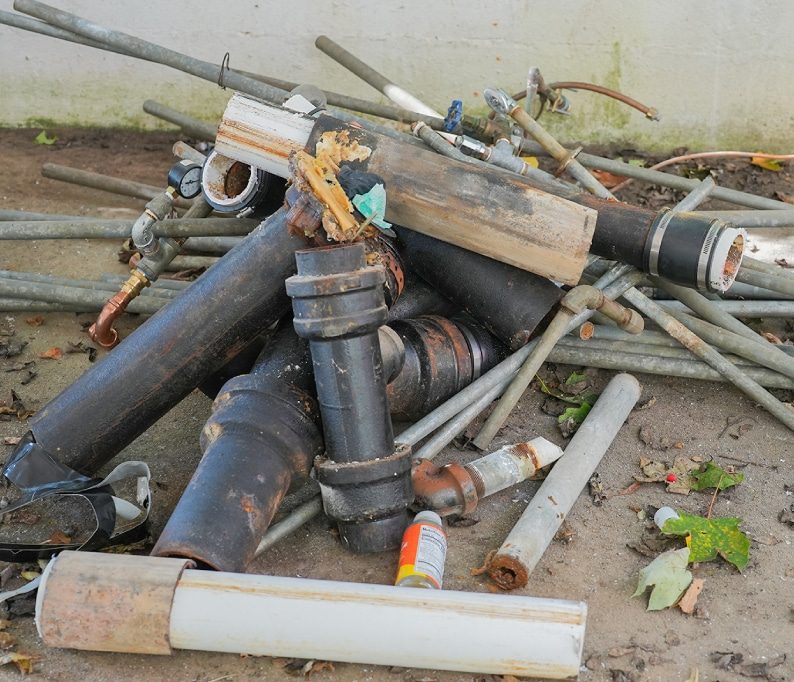 A pile of used black plastic and metal pipes, fittings, and debris lying on the ground near a wall.