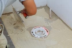 A person spreads tile adhesive with a notched trowel on a bathroom floor around a shower drain in preparation for tiling.