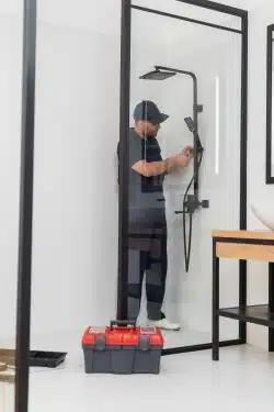 plumber in overalls repairing shower in bathroom near toolbox and tools on floor
