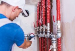A plumber in a blue shirt adjusts a pressure gauge on a pipe system with multiple red insulated pipes and valve handles, ensuring thorough plumbing inspections for Oak Ridge.