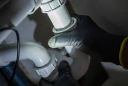 A person wearing dark gloves is installing a kitchen sink drain, tightening a white plastic pipe under a sink and focusing light on the plumbing connection.