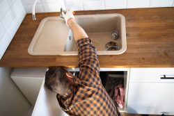 A person wearing a plaid shirt and glove repairs a kitchen faucet during a Plumbing Renovation in Knoxville, reaching under the sink on a wooden countertop.
