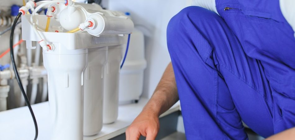 A person in blue overalls is installing or servicing a reverse osmosis water filtration system under a kitchen sink, highlighting the importance of professional Water Treatment Systems in Oak Ridge.