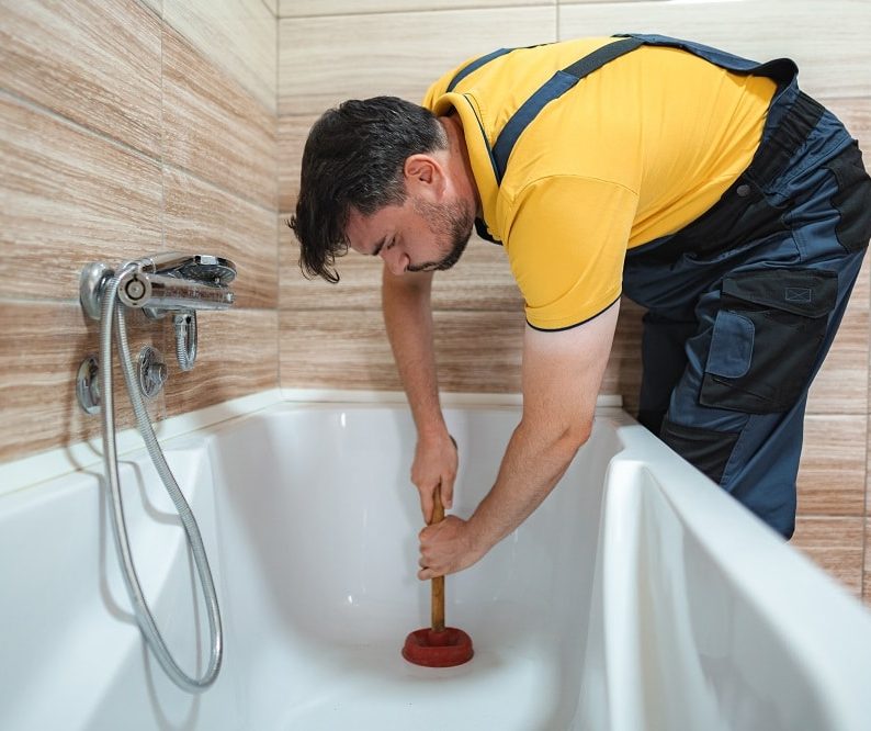 Person wearing gloves uses a wrench to tighten a bolt on a metal fixture beneath a white bathtub, possibly working to unclog a bathtub drain, all set on a wooden floor.