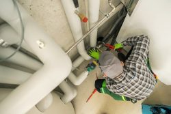 A plumber wearing a cap and gloves uses a wrench to work on pipes in a utility room, ensuring top-quality plumbing inspections for Oak Ridge.