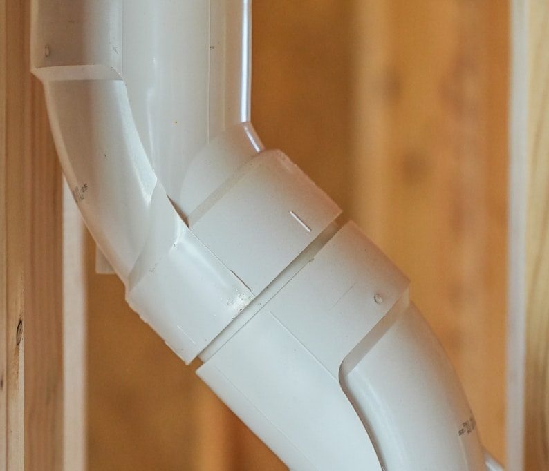 Close-up of a white PVC pipe joint with a bend, installed within a wooden framing structure.