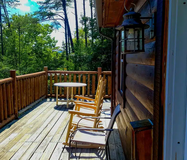 Wooden deck with three chairs and a round table outside a log cabin, surrounded by trees under a partly cloudy sky.