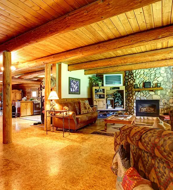 A cozy log cabin interior with exposed wooden beams, stone fireplace, patterned sofas, and warm ambient lighting.