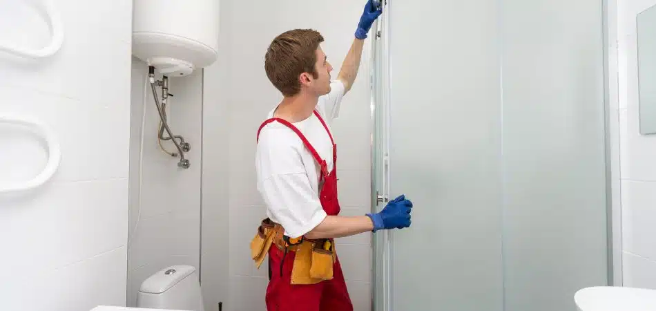 A repairman in red overalls and blue gloves installing or adjusting a glass shower door in a bright white bathroom, with a tool belt around his waist.